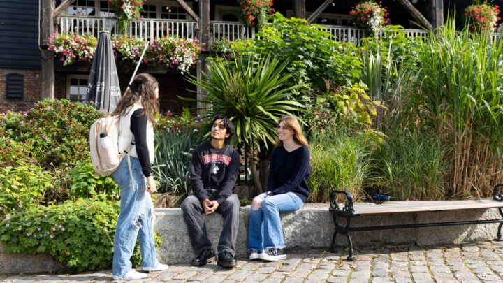Three young people talk outdoors by a stone bench surrounded by lush greenery and flowers, with a building and balcony decorated with flower boxes in the background.