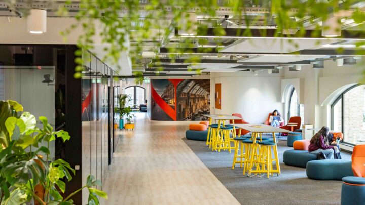 A modern, open office space with plants, lounge seating, high tables with yellow stools, and people working. Large windows and bright decor create a welcoming, collaborative atmosphere.