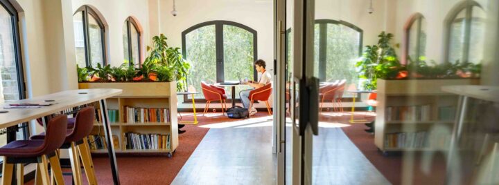 A person sits at a table by large arched windows in a bright, modern library or study area with bookshelves, plants, and red chairs along the wall. Their reflection is visible on a glass wall.