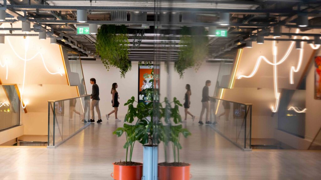 Four people walk across a modern, brightly lit indoor walkway with glass railings, hanging plants, and a polished floor, which reflects the scene. Contemporary decor and neon-style lighting are visible.