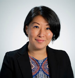 Chong Kim-Wong, with short, dark hair, smiles while wearing a black blazer over a blue-patterned shirt. The background is a simple, light gradient.
