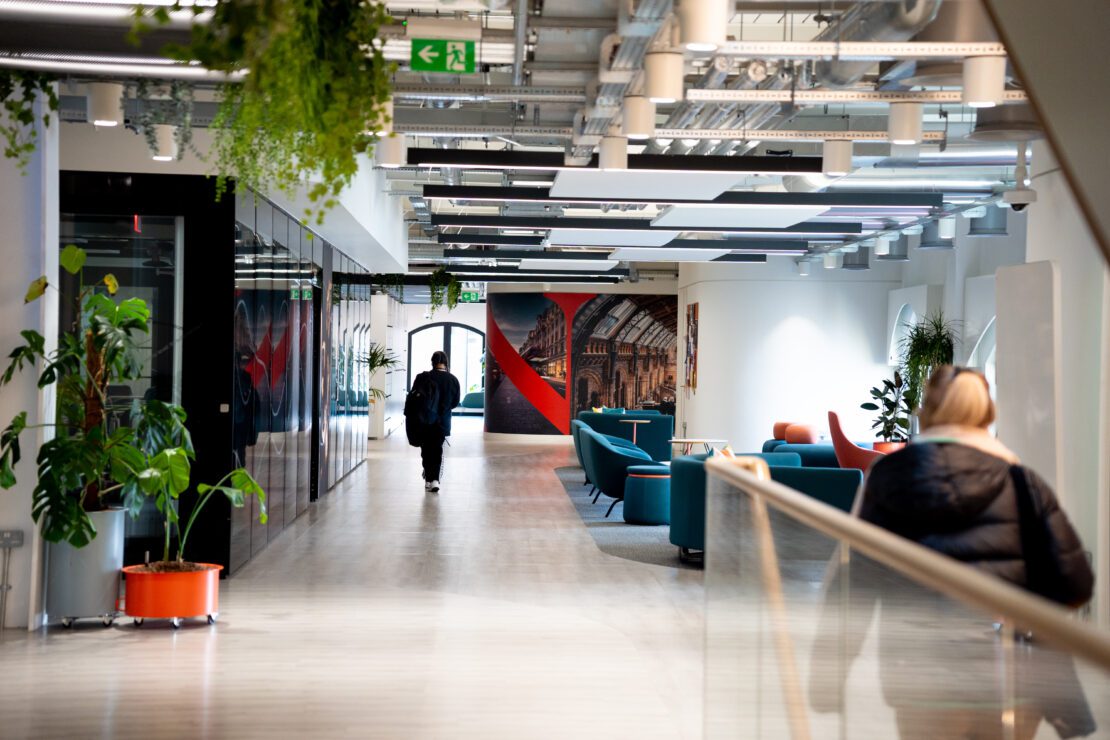 A modern office hallway with a person walking and another standing at the railing. The space features plants, a seating area with teal chairs, and a wall mural. Ceiling pipes and ducts are visible, with ample natural light flooding the area.