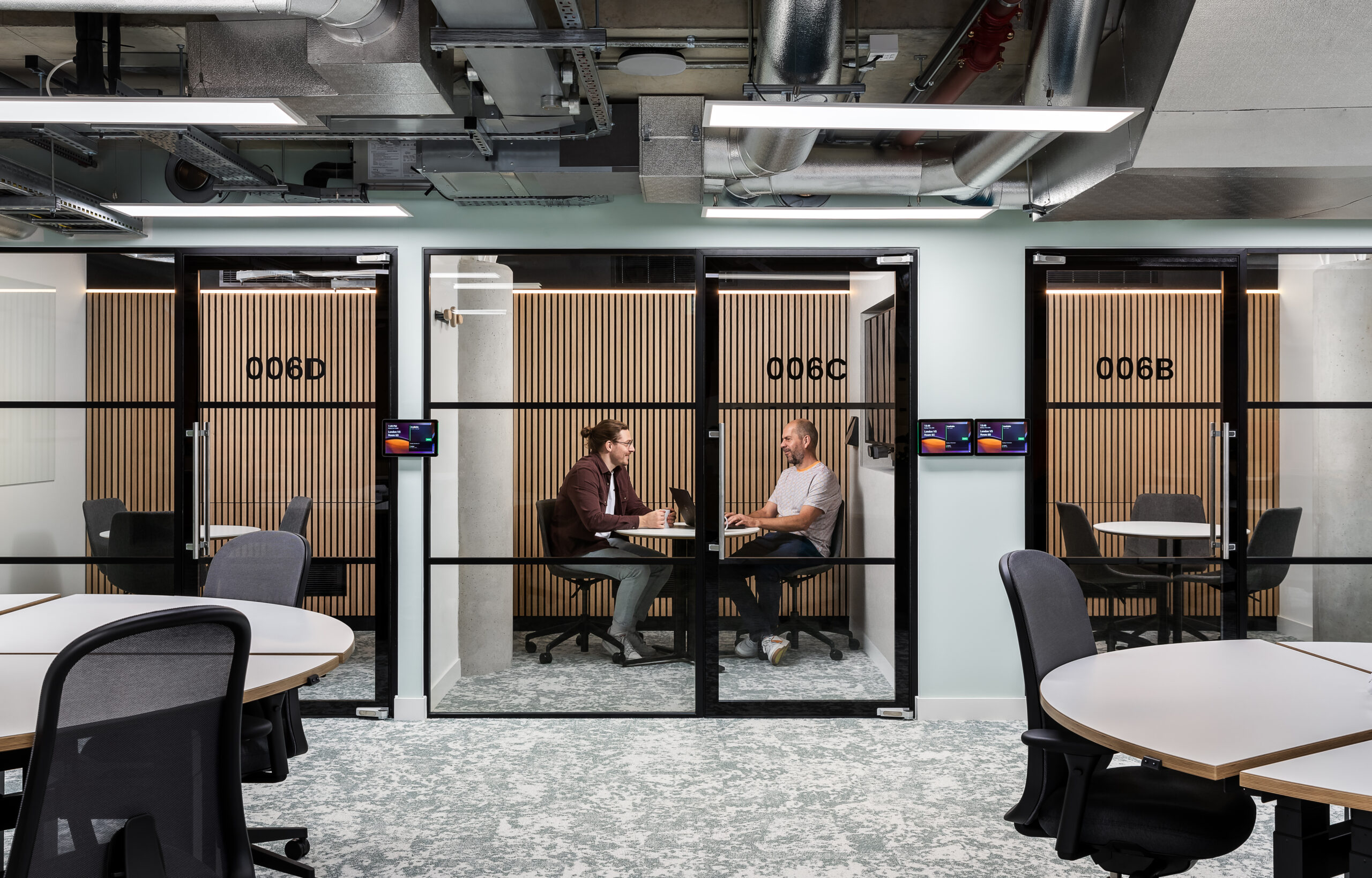 Two people are sitting at a small table in a glass-walled meeting room labeled "006B." The room features wooden panels and digital screens outside. The surroundings include modern office furniture and exposed ceiling pipes.