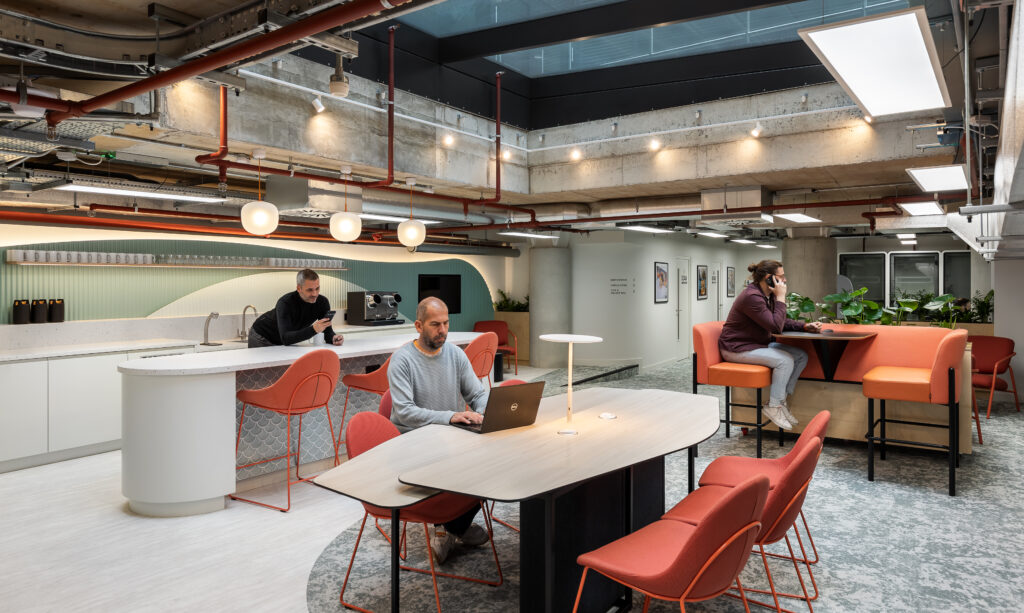 A modern office space with three people working. One person is seated at a table with a laptop, another is using a phone at a booth, and the third is making coffee. The room has orange chairs and overhead lighting.