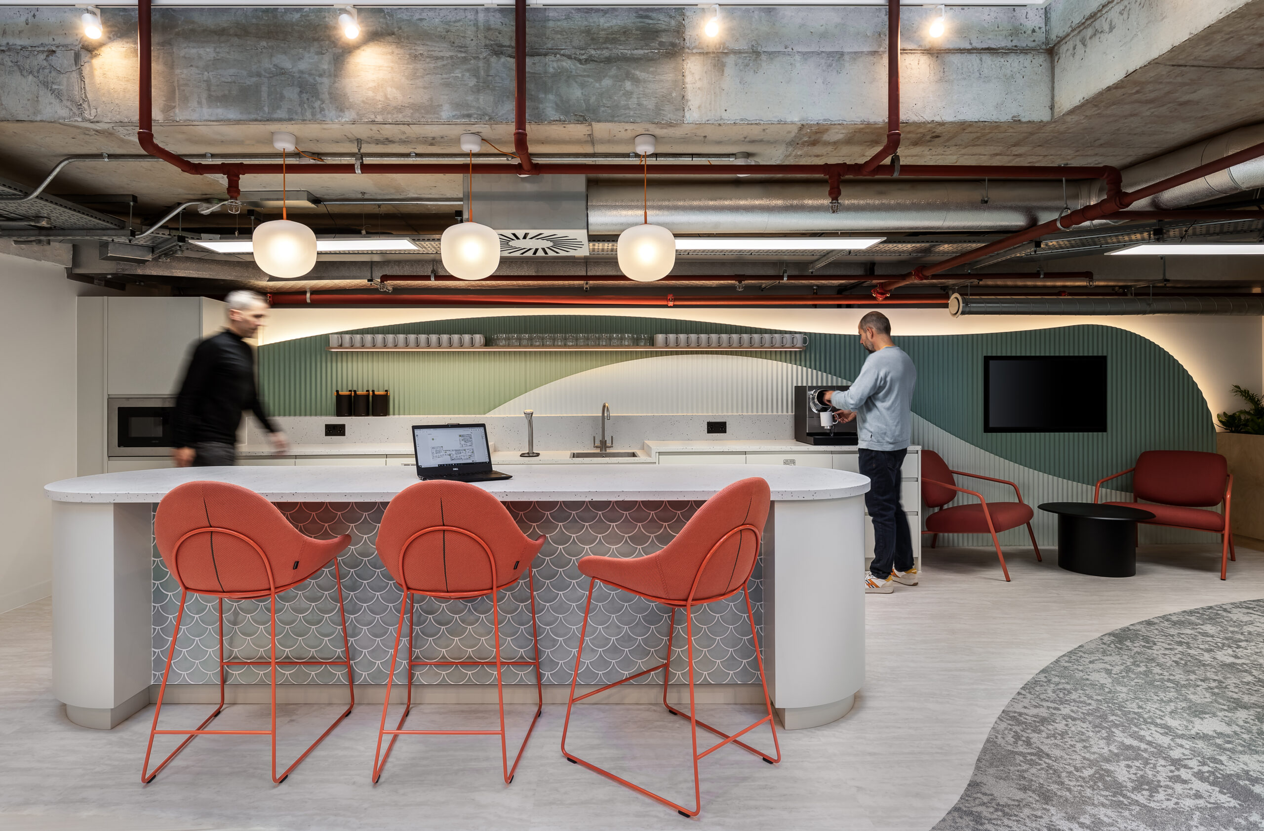 A modern office break area with a sleek kitchen counter, three orange chairs, and a laptop. Two people interact in the space. The design features exposed pipes, pendant lighting, and a mix of textures and colors, including red and green accents.
