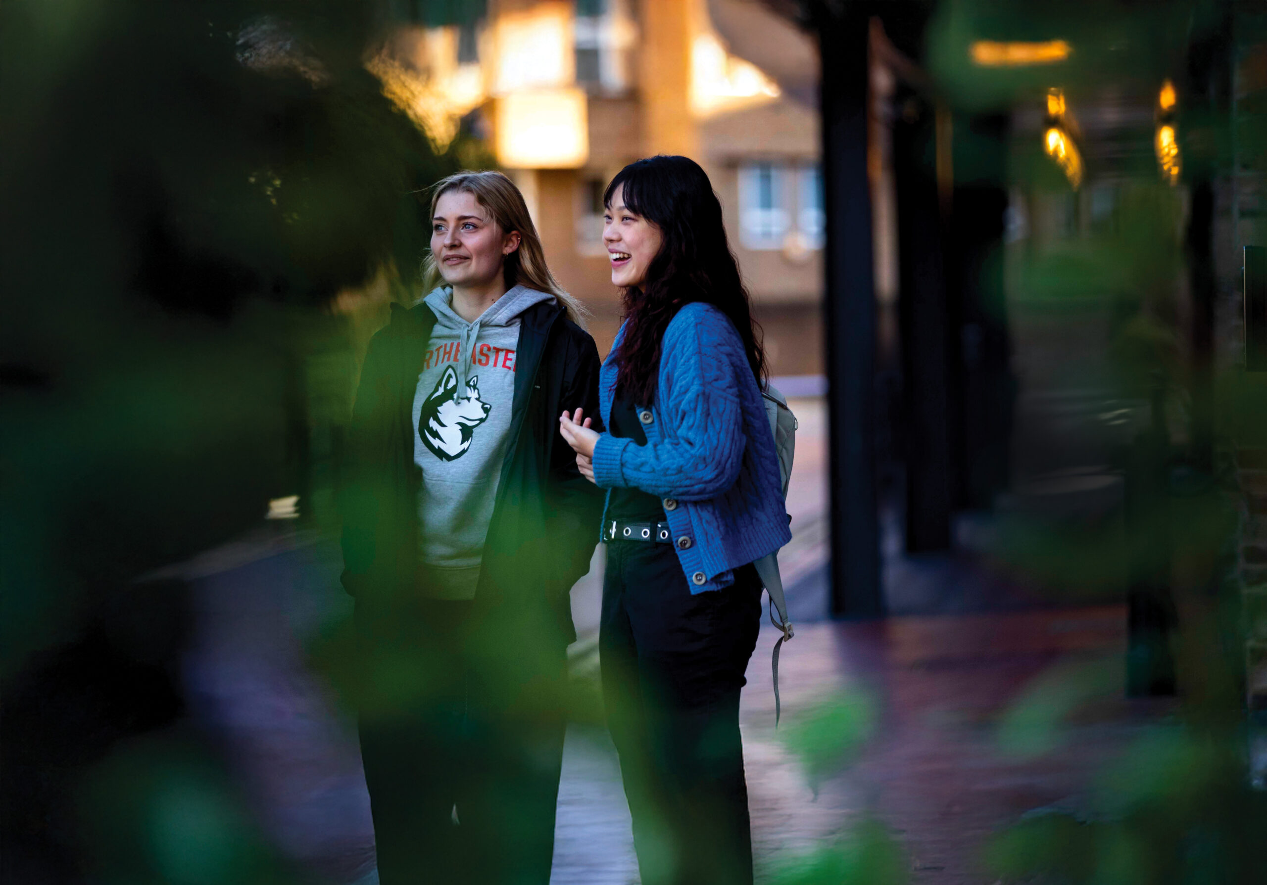 Two people walk together on a brick path, partially obscured by green leaves. One wears a gray hoodie, and the other wears a blue sweater. They're smiling and engaged in conversation, with buildings softly blurred in the background.