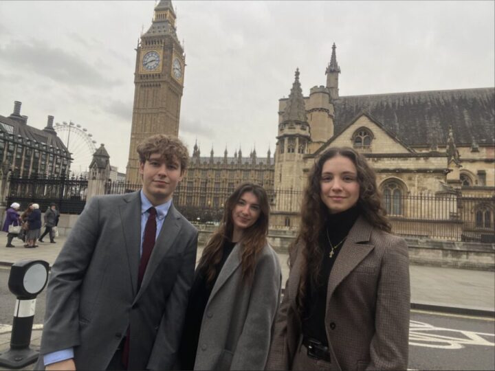 Three people in business attire stand in front of the Houses of Parliament in London. Big Ben and the London Eye are visible in the background. The sky is overcast.