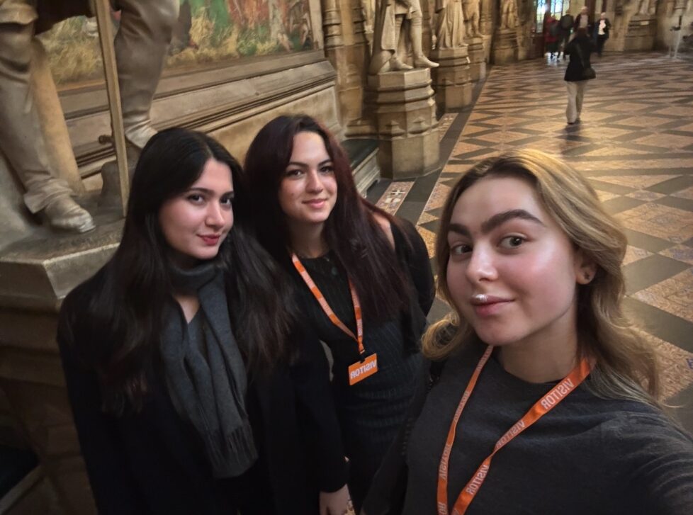 Three women wearing visitor passes stand in a grand hall with elaborately carved statues and a patterned floor. They are smiling and posing for a selfie.