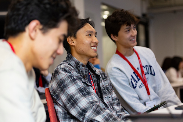 Three young adults are sitting in a classroom, wearing casual clothes and red lanyards. They are smiling and engaged, looking towards the front of the room. The setting is lively, and there are more people blurred in the background.