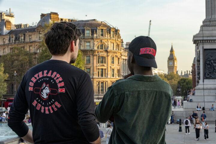 Two people wearing Northeastern Huskies apparel look towards Big Ben in London. The setting is urban with historical architecture, and several people are visible in the background enjoying the day.
