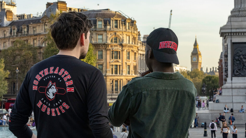Two people wearing Northeastern Huskies apparel look towards Big Ben in London. The setting is urban with historical architecture, and several people are visible in the background enjoying the day.