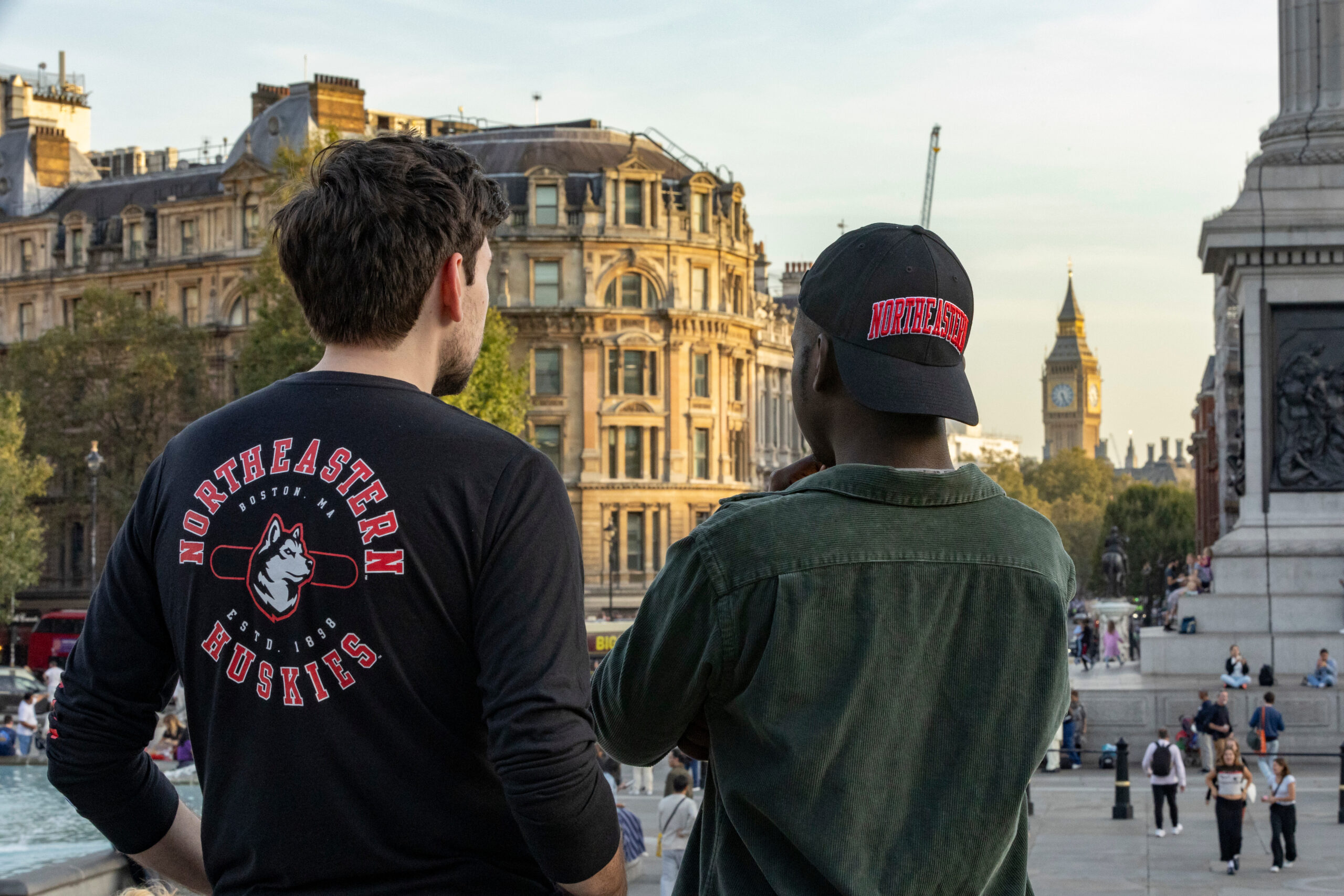 Two people wearing Northeastern Huskies apparel look towards Big Ben in London. The setting is urban with historical architecture, and several people are visible in the background enjoying the day.