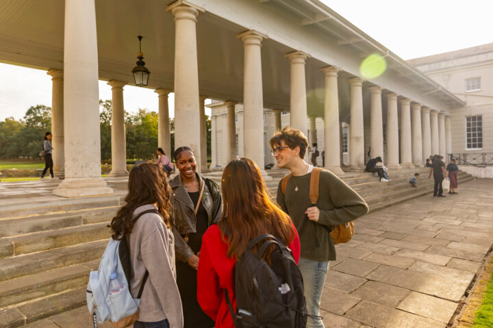 Four people with backpacks and casual clothing stand talking in front of a neoclassical building with tall columns. The sun is shining, and a few others are visible in the background, enjoying the outdoor setting.