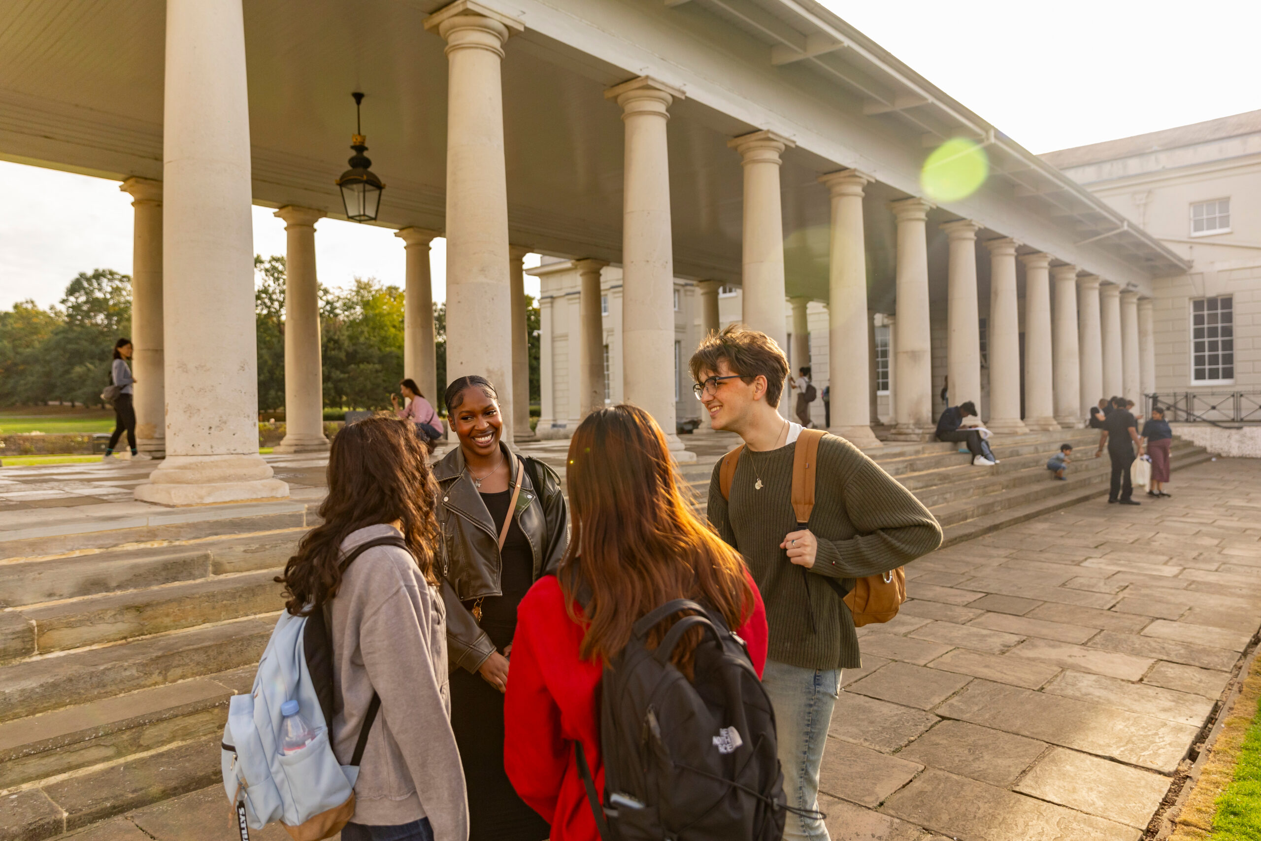 Four people with backpacks and casual clothing stand talking in front of a neoclassical building with tall columns. The sun is shining, and a few others are visible in the background, enjoying the outdoor setting.