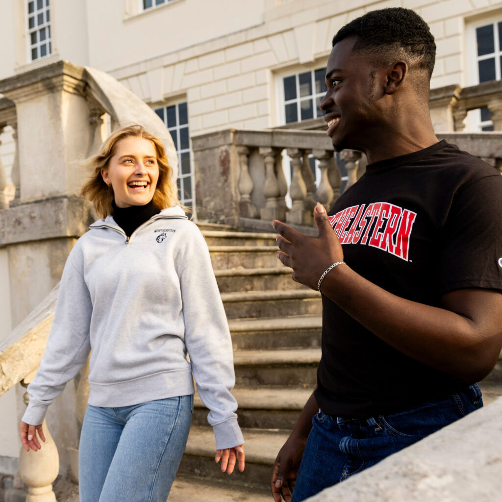 A woman in a gray sweatshirt and jeans and a man in a black T-shirt are walking down stone steps outside a building. Both are smiling and engaged in conversation. The setting is bright and sunny.