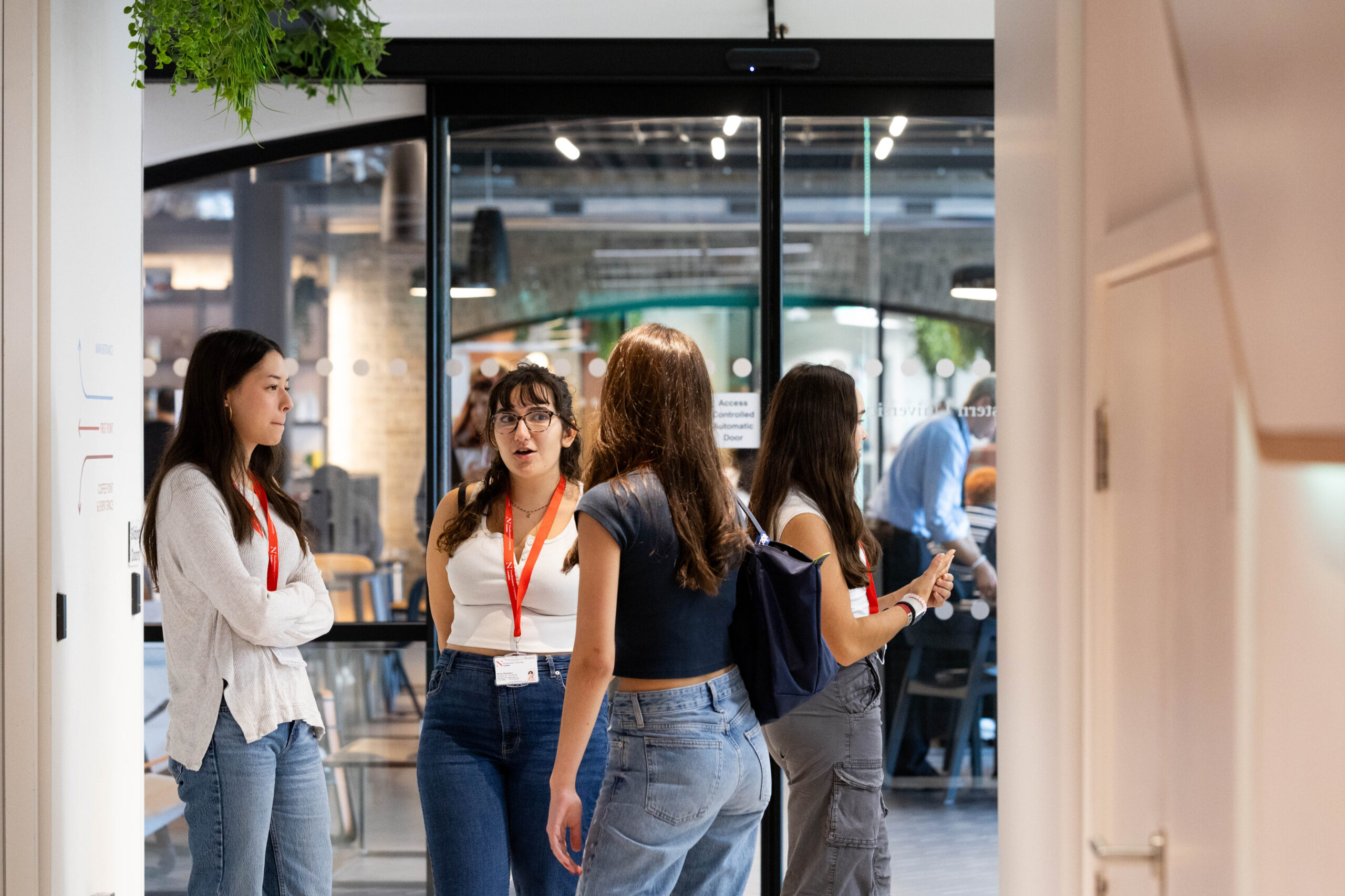 Four people wearing lanyards stand in a group, talking in a modern indoor space. Some have shoulder bags and are casually dressed. There's a glass wall and a hanging plant above them. More people and tables are visible in the background.