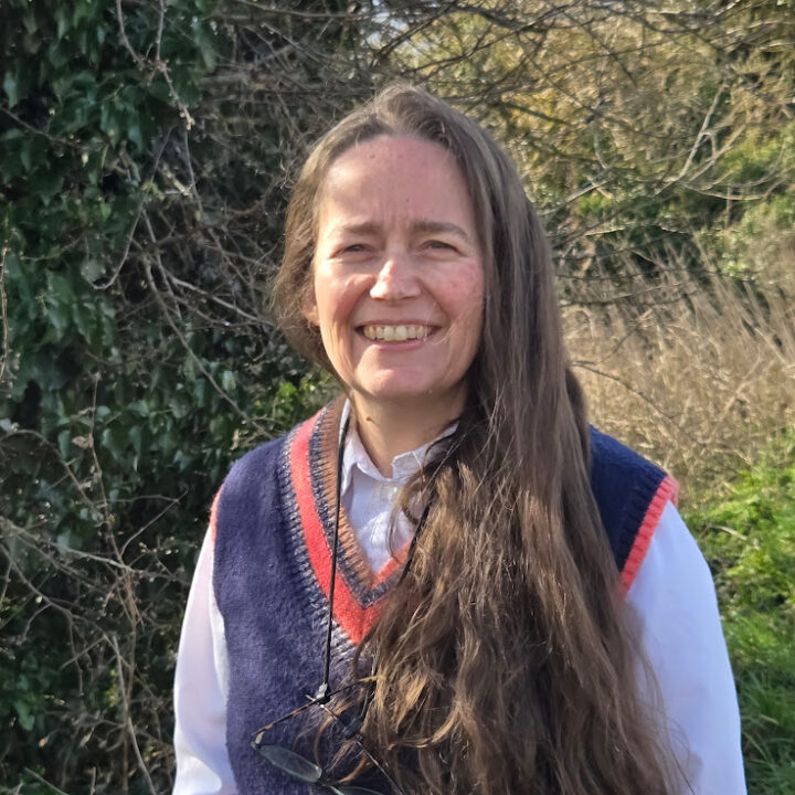 Smiling woman with long hair wearing a white shirt and a blue vest stands outdoors. Green foliage and leafless branches are visible in the background.