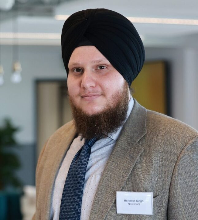 A person with a beard and wearing a turban is dressed in a suit and tie. They have a name tag, and stand indoors with blurred background elements like doors and lights.