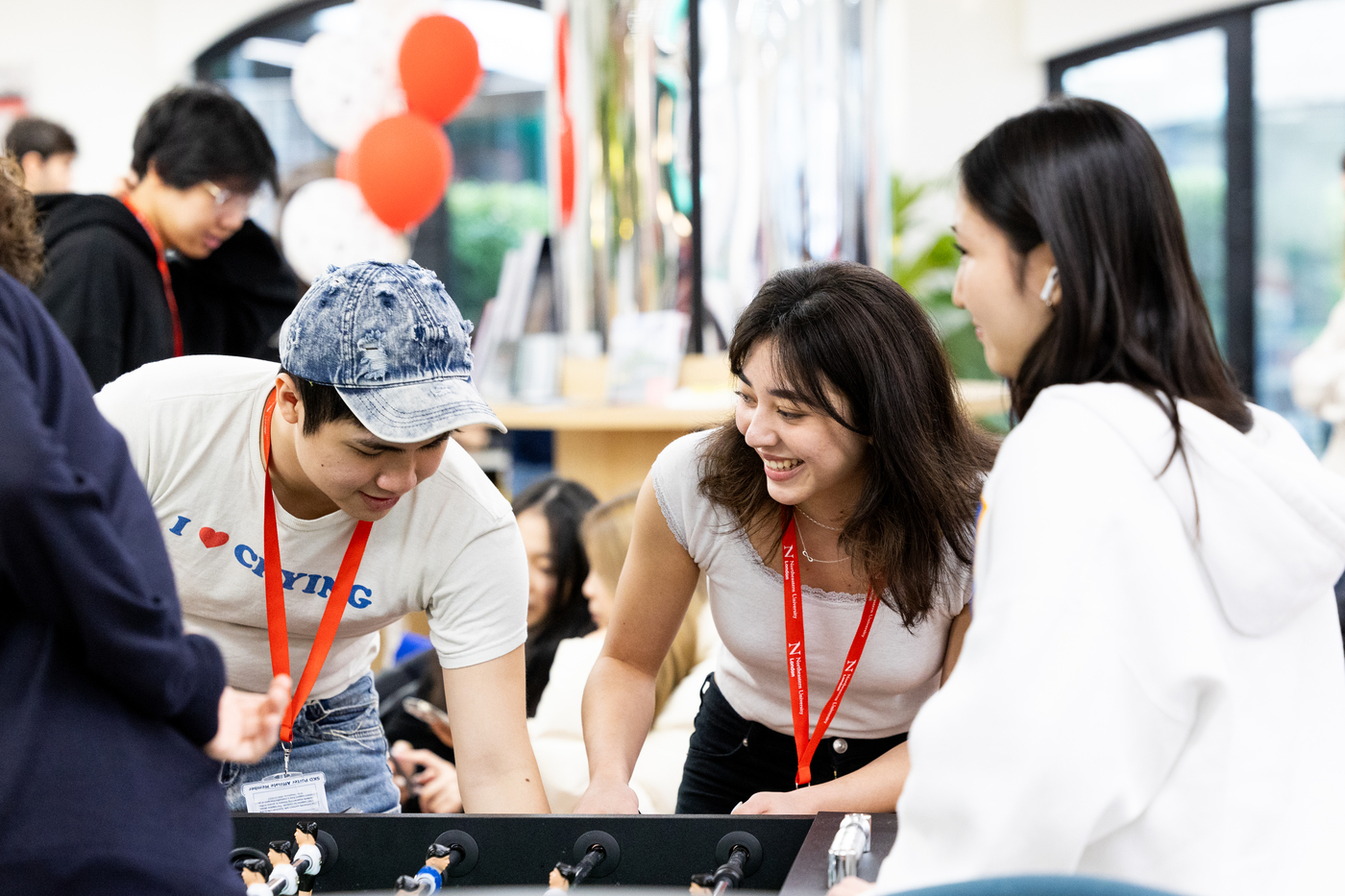 Three people enjoy a foosball game in a lively indoor setting. Two women smile while playing, and a man in a cap studies the game. Balloons and other people are visible in the blurred background.