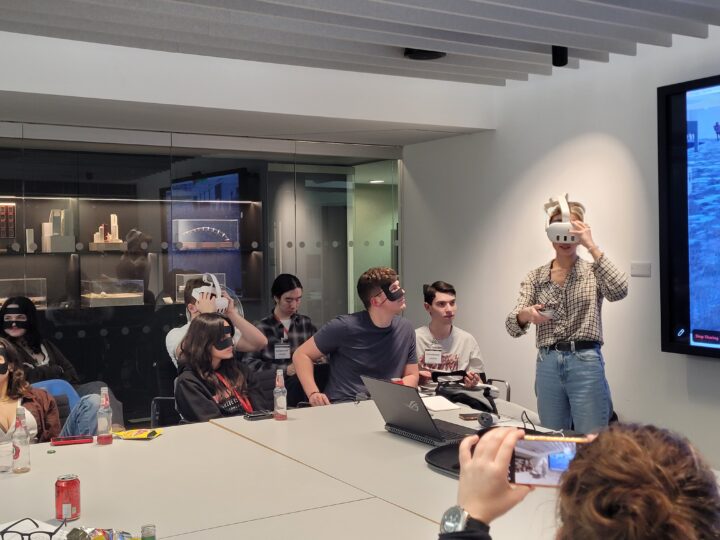 A group of people sit around a conference table, some wearing virtual reality headsets. One person stands and gestures while wearing a headset. A large screen and laptops are visible in the modern meeting room.