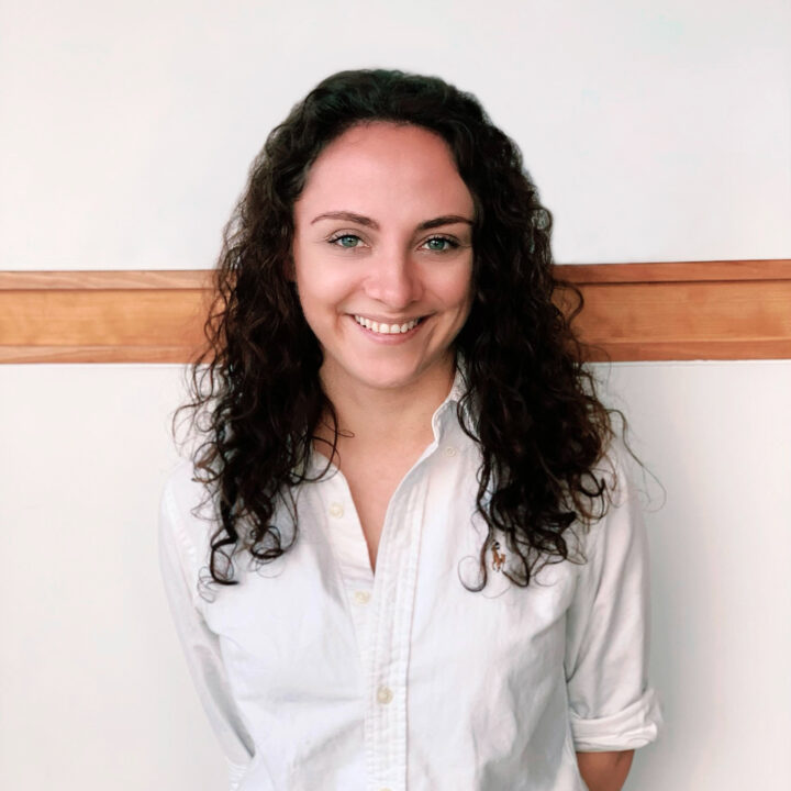 A woman with curly brown hair, wearing a white button-up shirt, stands smiling in front of a white wall with a wooden horizontal trim.