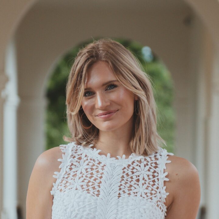 A woman with shoulder-length blonde hair smiles while wearing a sleeveless white lace top, standing outdoors in front of an archway with greenery in the background.