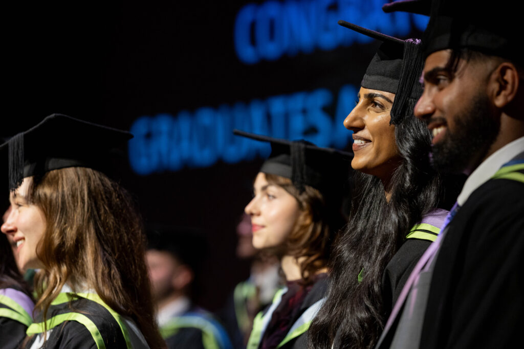 A group of graduates in caps and gowns smile as they stand in a row at a graduation ceremony, with congratulatory words blurred in the background.