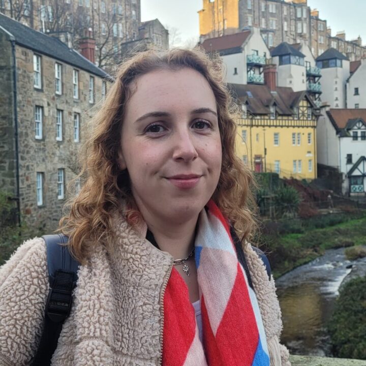 A young woman with curly hair and a colorful scarf smiles gently outdoors, standing in front of old stone buildings and yellow houses, with trees and taller buildings in the background.
