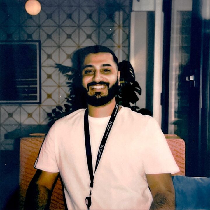 A man with a beard and short hair smiles while sitting indoors on a patterned chair. He is wearing a white t-shirt and a black lanyard, with plants and geometric-patterned walls in the background.