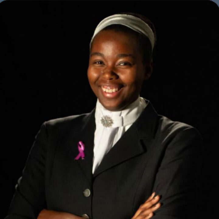 A woman smiles with arms crossed, wearing a black blazer, white shirt, and headband. She has a pink ribbon pin and a decorative brooch on her collar, standing against a dark background.