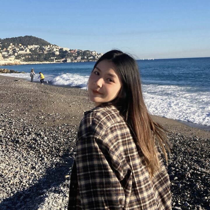 A young woman with long hair in a plaid shirt stands on a rocky beach, looking back at the camera. The ocean waves crash nearby under a clear blue sky, with hills and buildings visible in the background.