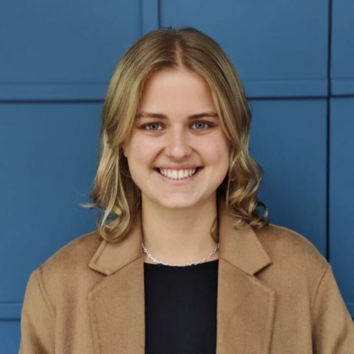 A young woman with blonde hair, wearing a tan coat over a black top, smiles in front of a blue geometric-patterned wall.