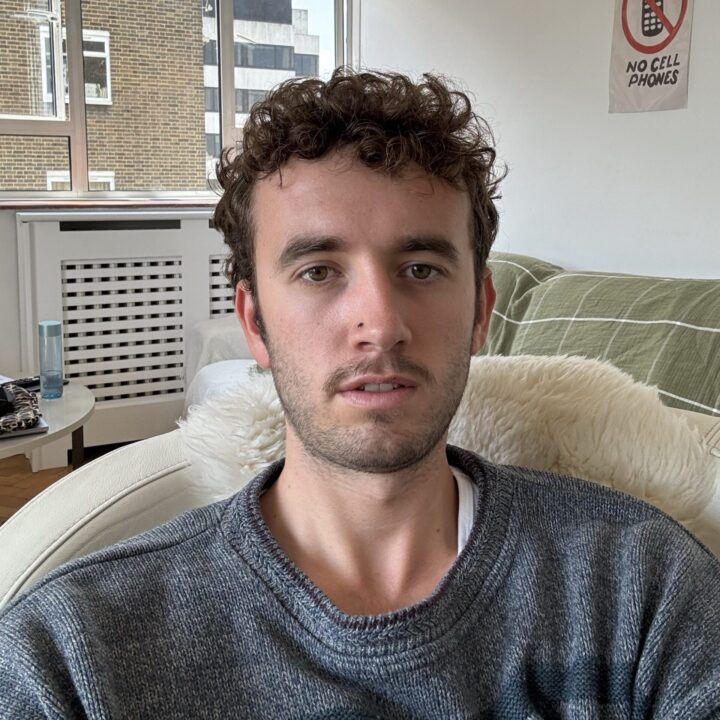 A young man with curly hair and a grey sweater sits on a couch in a bright room with large windows. A “No Cell Phones” sign is visible on the wall behind him.