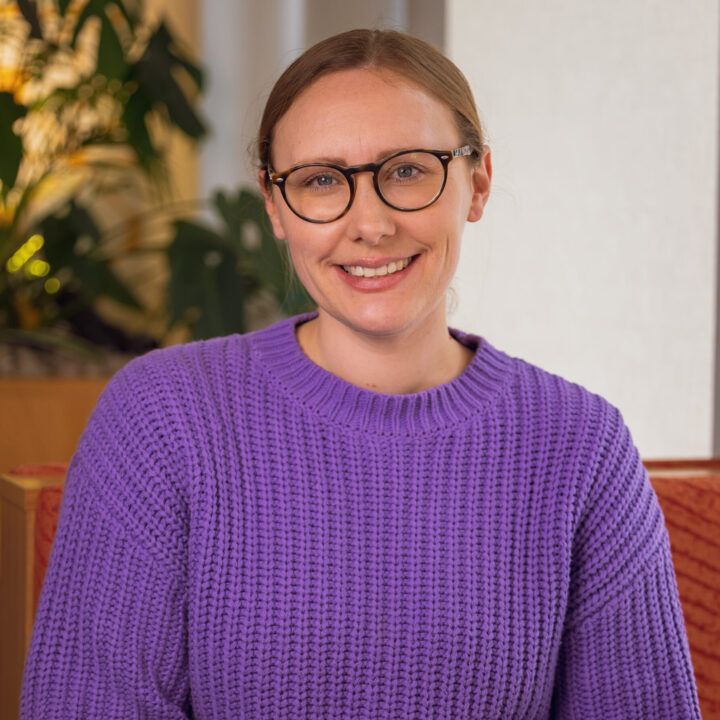 A woman with glasses and light brown hair tied back is smiling while sitting at a table. She is wearing a textured purple sweater and dark pants. The background features plants and soft, warm lighting.