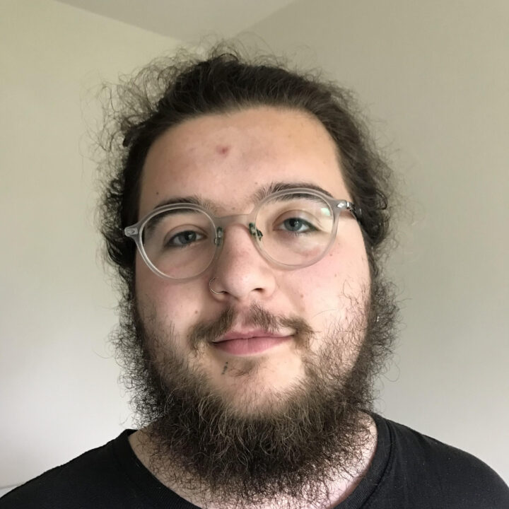 A person with long brown hair, a full beard, and glasses is standing indoors against a plain light-colored wall, wearing a black shirt and looking at the camera with a slight smile.