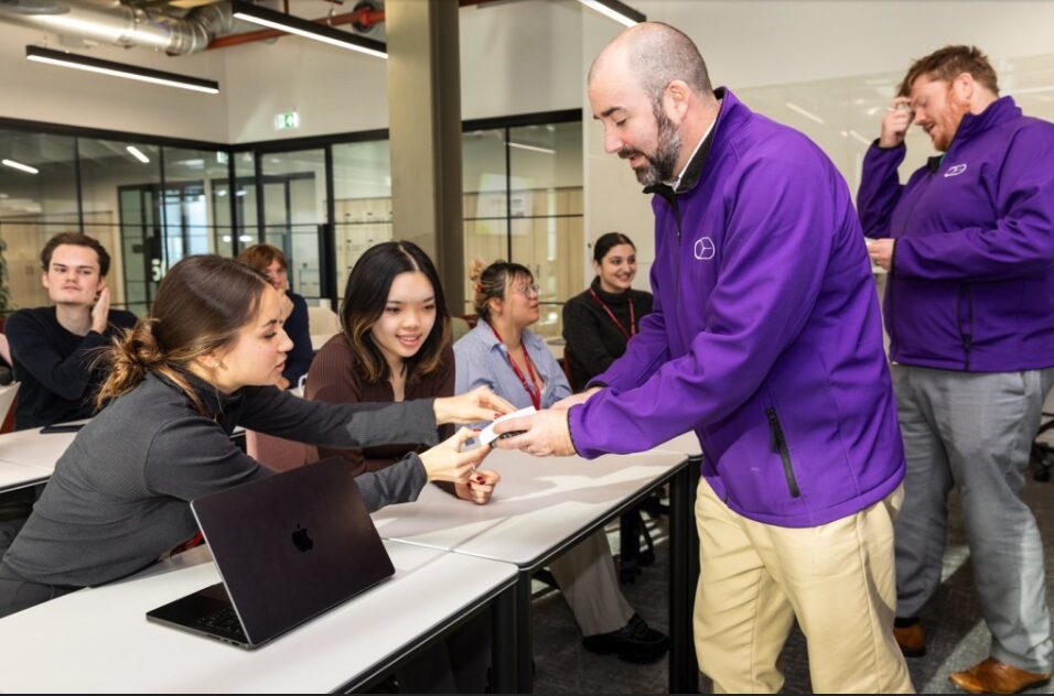 A person in a purple jacket hands a device to a seated person in a classroom setting. Other people sit and stand nearby, with laptops on desks. The room has glass walls and industrial-style decor.