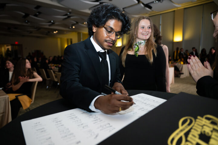 A man in a suit signs a large white document at a formal event, while people in formal attire look on in a well-lit room.