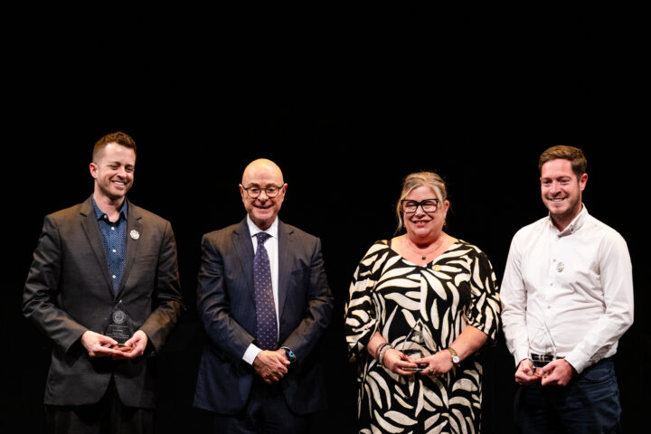 Four people stand side by side against a black background, three holding glass awards and smiling. The group includes two men in suits, a woman in a patterned dress, and a man in a white shirt.