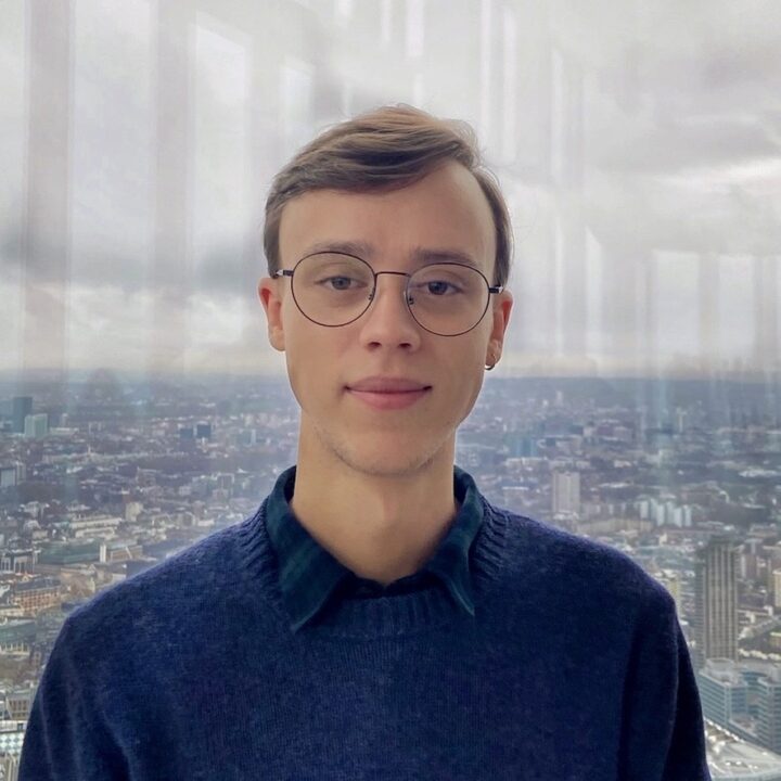 A young person with glasses and short light brown hair, wearing a blue sweater over a collared shirt, stands indoors in front of a large window overlooking a cityscape on a cloudy day.