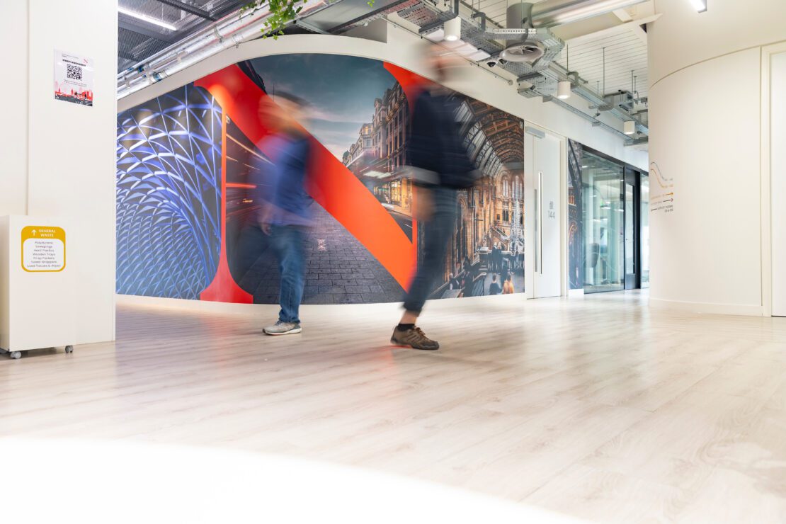 Two people walk through a modern, brightly lit hallway with a colorful mural featuring architectural images and a large red "N" on the curved wall. The floor is light wood, and the ceiling shows exposed pipes.