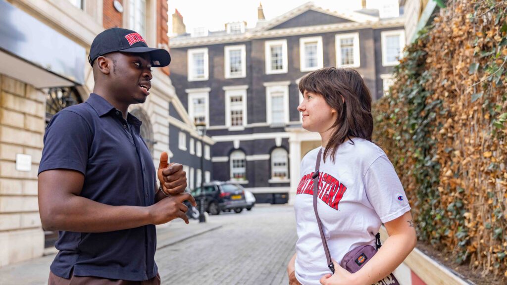 A man and a woman stand on a cobblestone street, engaged in conversation. The man wears a black cap and shirt, while the woman wears a white t-shirt and carries a small bag. Townhouses and parked cars are in the background.