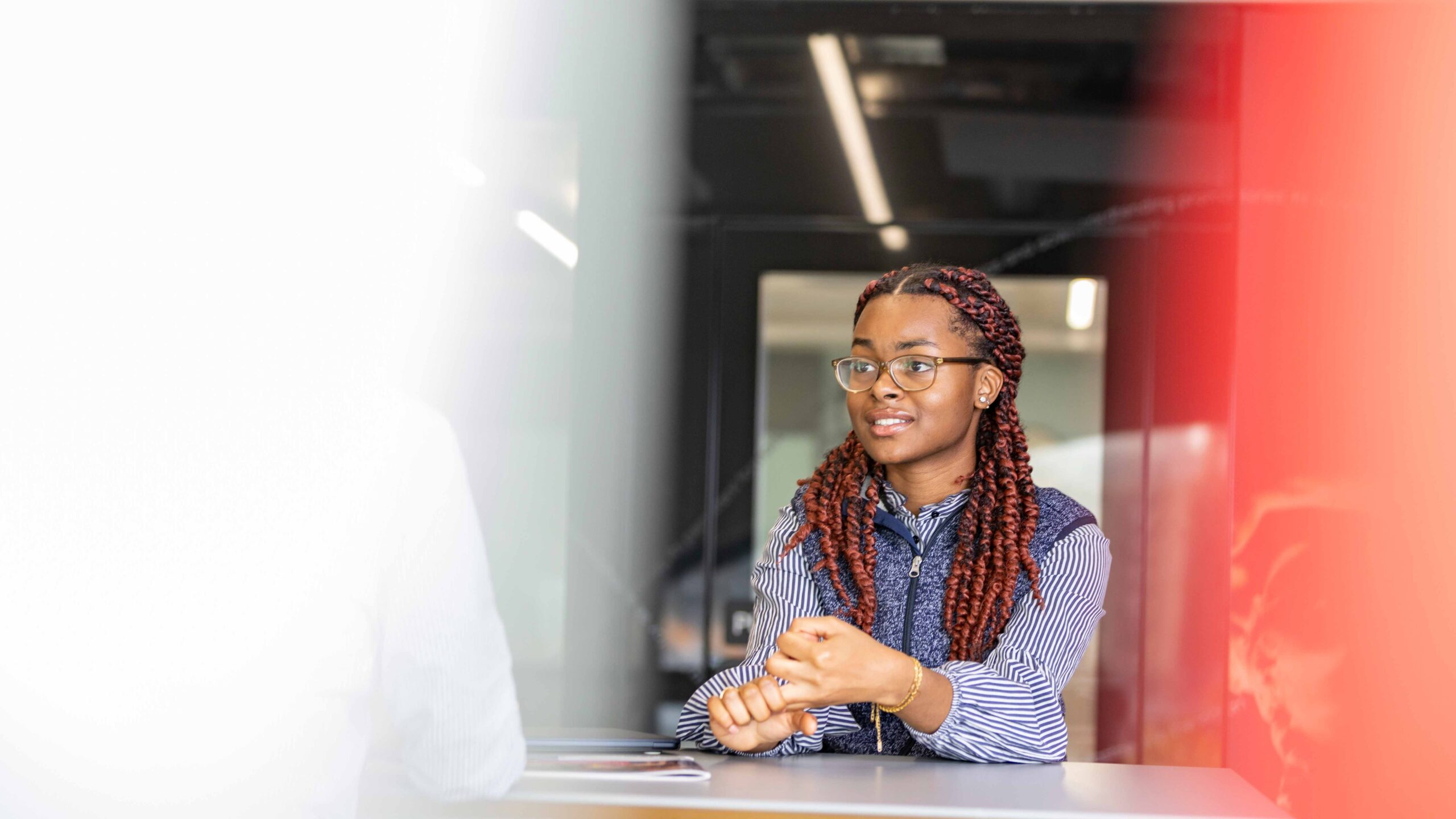 A woman with long braided hair and glasses sits at a table indoors, engaged in conversation. She wears a striped shirt and gestures with her hands. The background is blurred with red and white hues framing the image.