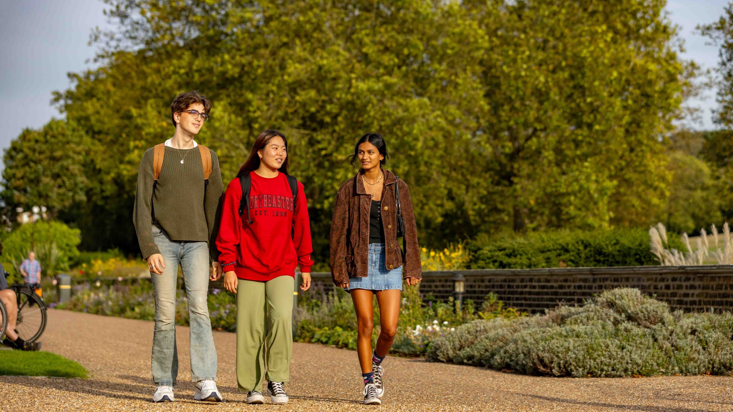 Three young adults walk together on a path in a park, surrounded by greenery and trees. They are casually dressed and smiling, enjoying a sunny day outdoors.