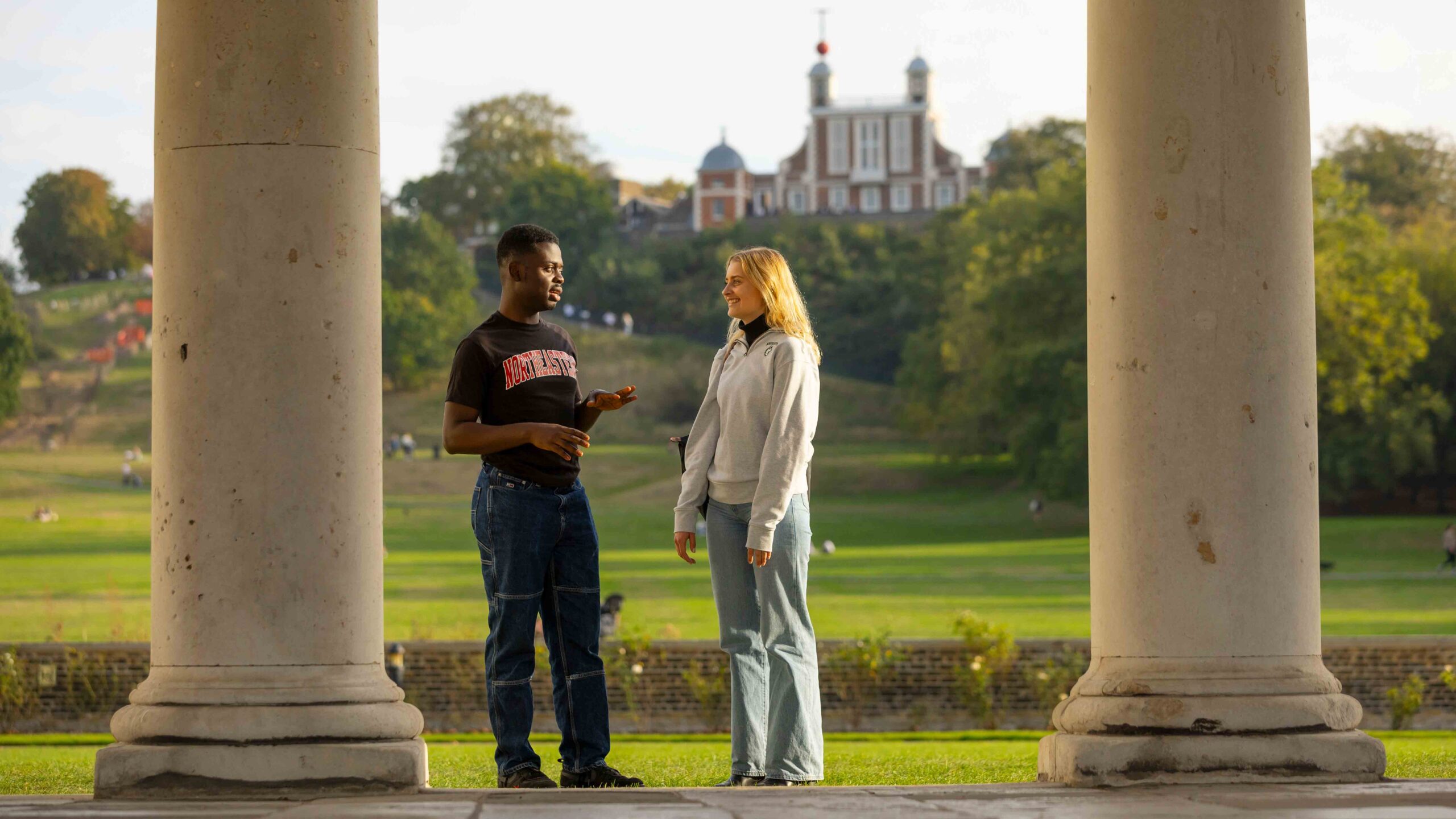 Two people stand and talk between large stone columns in a park, with green trees and a historic building on a hill visible in the background.