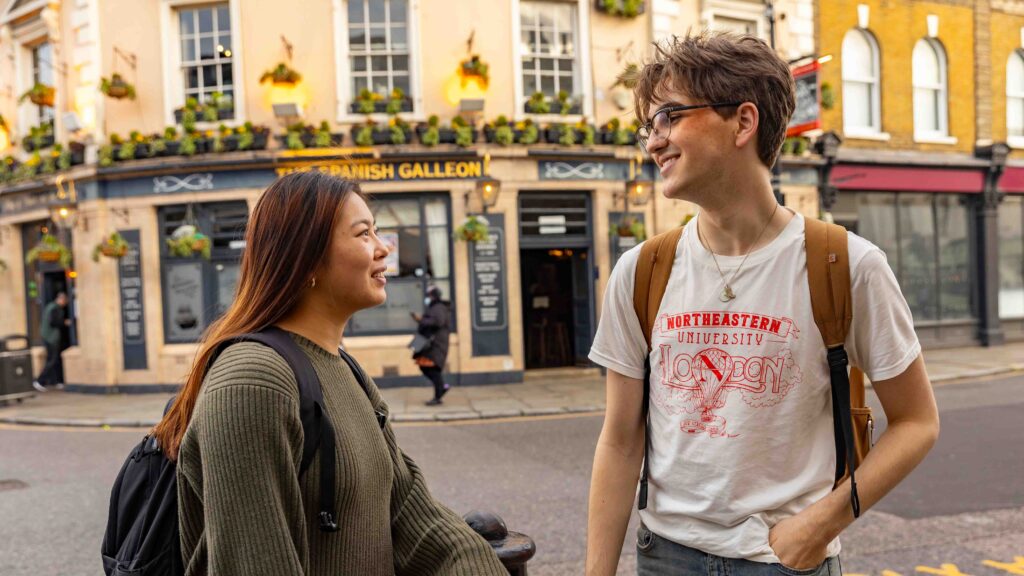 Two young adults with backpacks smile and talk while standing on a street corner in front of a building with flower boxes and a sign that reads "The Spanish Galleon.
