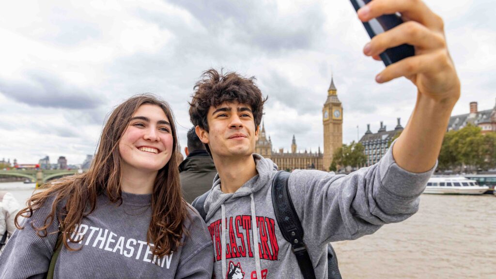 Two young people wearing Northeastern University sweatshirts take a selfie by the River Thames, with Big Ben and the Houses of Parliament visible in the background under a cloudy sky.