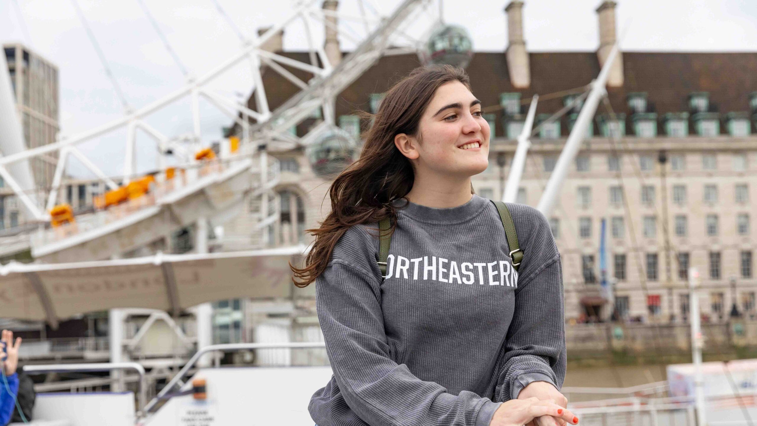A smiling young woman wearing a Northeastern sweatshirt stands outdoors in front of an observation wheel and historic buildings, with a river and cityscape in the background.