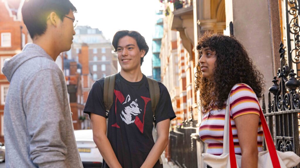 Three young adults stand outside on a city street, talking and smiling. Two men and one woman, all casually dressed, are engaged in conversation near a black iron fence and brick buildings.