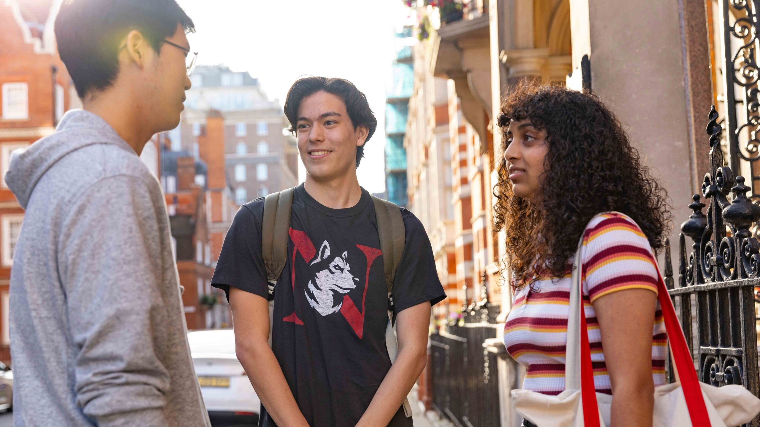 Three young adults stand outside on a city street, talking and smiling. Two men and one woman, all casually dressed, are engaged in conversation near a black iron fence and brick buildings.