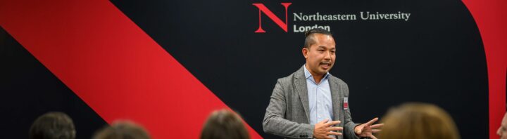 A man speaks to an audience in front of a wall with the Northeastern University London logo and red and black diagonal stripes.